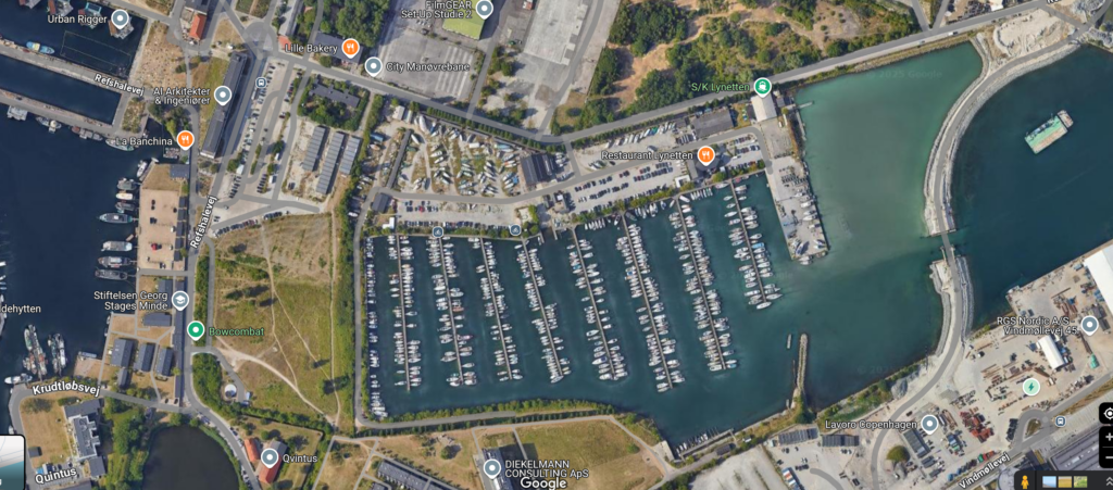 Luftfoto af en marina med talrige både, der ligger til kaj i en havn. De omkringliggende områder omfatter veje, bygninger og græsklædte pletter. Vandet skaber et naturligt indløb, og parkeringspladser er synlige.